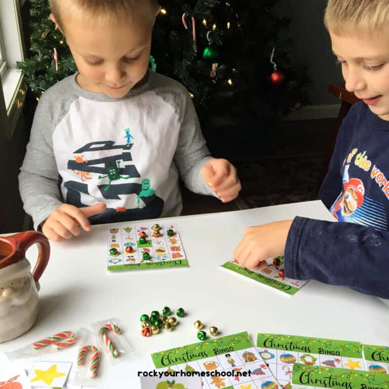 Two young boys playing Christmas bingo game in front of Christmas tree with candy canes, Santa Claus mug, and jingle bells.