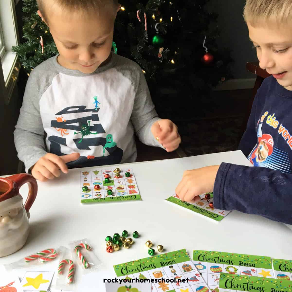 Two young boys playing Christmas bingo game in front of Christmas tree with candy canes, Santa Claus mug, and jingle bells.