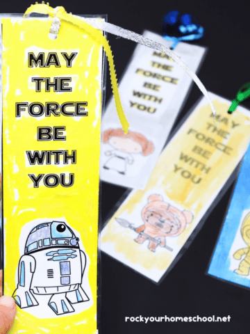Woman holding examples of Star Wars coloring bookmarks saying May The Force Be With You featuring R2-D2 with other bookmarks in the background.