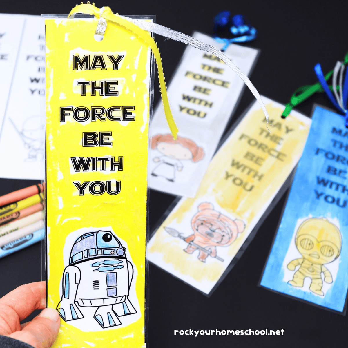 Woman holding examples of Star Wars coloring bookmarks saying May The Force Be With You featuring R2-D2 with other bookmarks in the background.