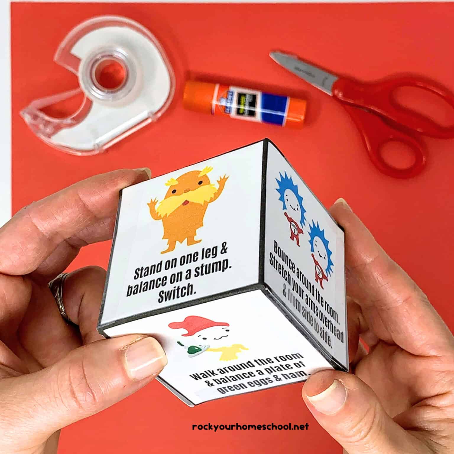 Woman holding Dr. Seuss-inspired activity cube featuring The Lorax, Thing 1, Thing 2, and The Fish with tape, glue stick, and scissors.