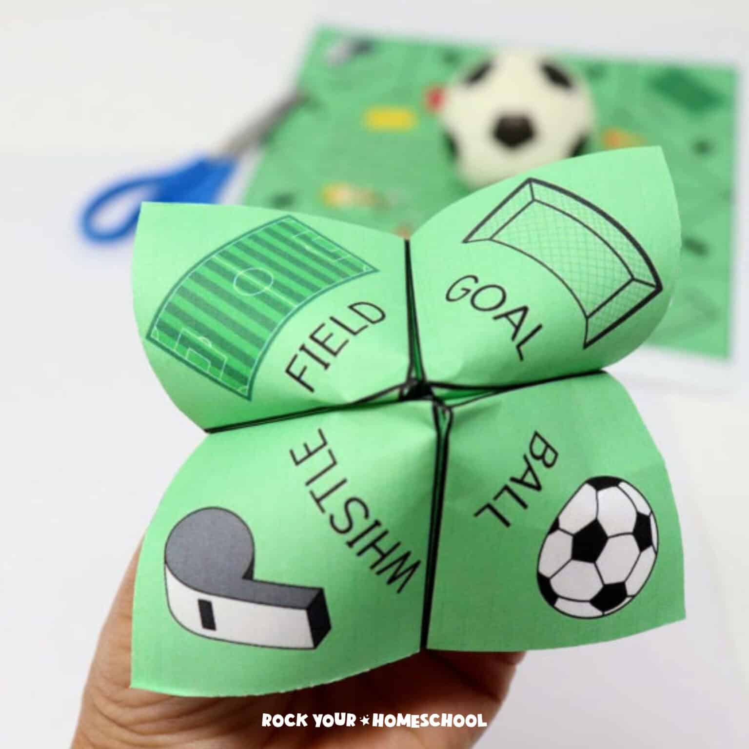 Woman's hand holding a soccer cootie catcher featuring a field, goal, whistle, and ball with blue scissors, soccer cootie catcher printable page, & small toy soccer ball in background.