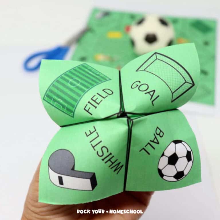 Woman's hand holding a soccer cootie catcher featuring a field, goal, whistle, and ball with blue scissors, soccer cootie catcher printable page, & small toy soccer ball in background.