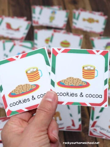 Woman holding pair of cookies & cocoa Christmas memory game cards with examples in background.