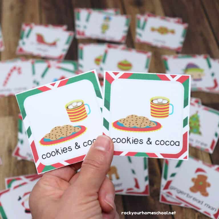 Woman holding pair of cookies & cocoa Christmas memory game cards with examples in background.