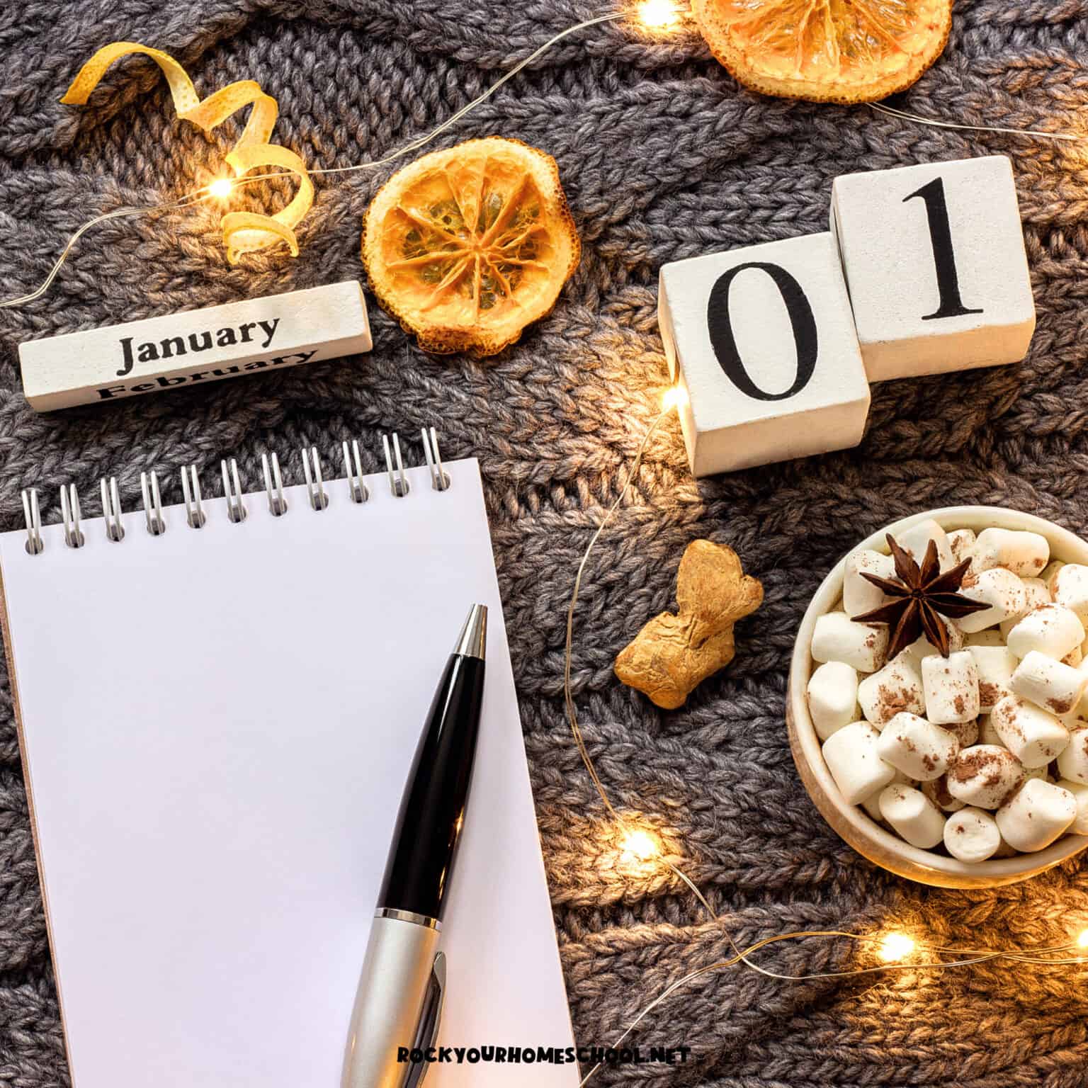 Notebook with pen, January, 01, dried orange slices, hot cocoa mug, and twinkle lights on gray knitted blanket to feature these January fun days for kids.