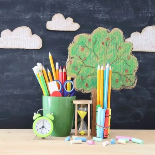 cardboard tree with color pencils and scissors in green mug with bright green alarm clock and sand time and colored chalk in front of blackboard with white cardboard clouds to feature homeschooling with Rock Your Homeschool
