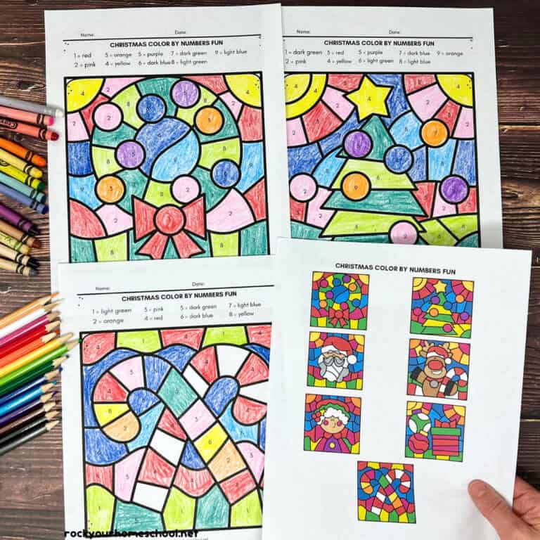 Woman holding answer key with examples of printable Christmas color by number pages with crayons and color pencils.