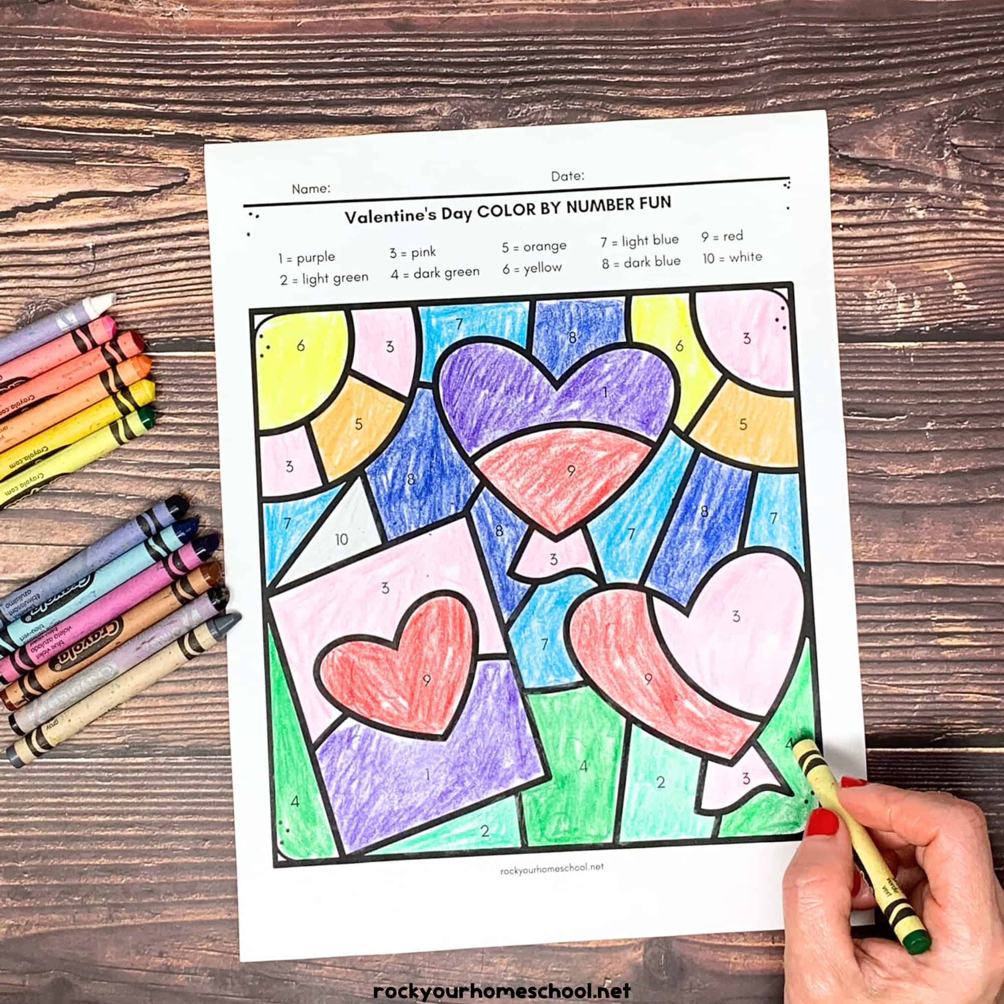 Woman holding green crayon and working on free printable Valentine's Day color by number page with heart balloons and card.