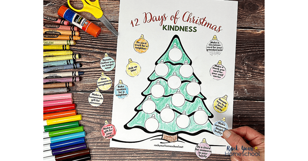 woman holding paper ornament with prompt in front of Christmas tree of 12 days of Christmas random acts of kindness with rainbow of markers, crayons, glue stick, and yellow stick on wood background