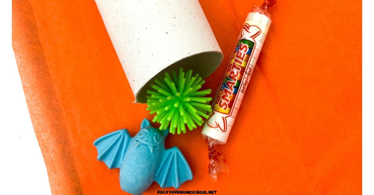 Toilet paper roll on orange tissue paper with Smarties candy, green plastic ball, an blue plastic bat.