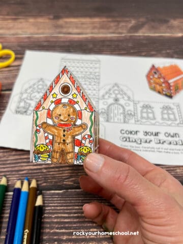 Woman holding example of 3D gingerbread house coloring craft with scissors, glue stick, and color pencils in background.