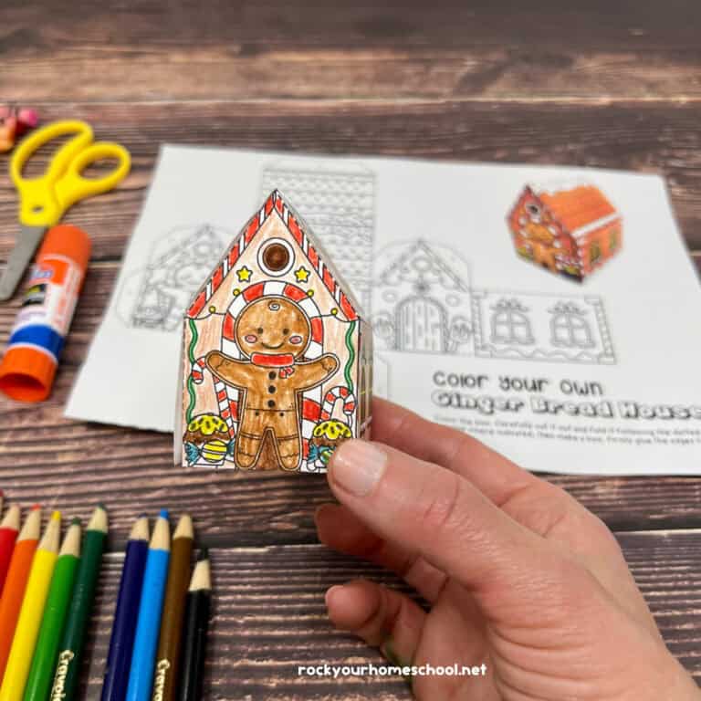 Woman holding example of 3D gingerbread house coloring craft with scissors, glue stick, and color pencils in background.