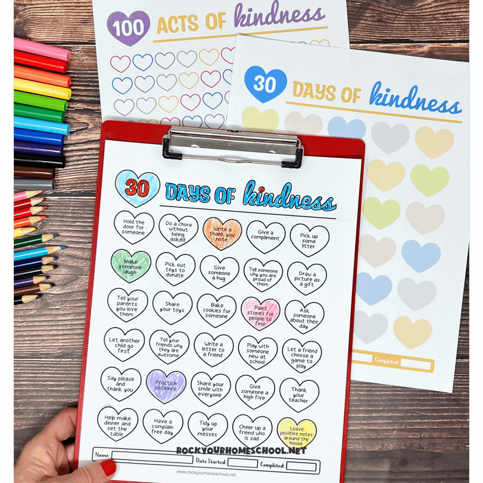 Examples of free printable kindness activities for kids on red clipboard and wood background with rainbow of markers and color pencils.