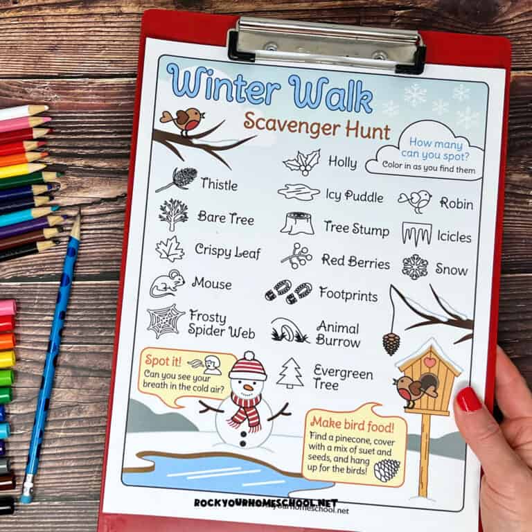 Woman holding red clipboard with free printable winter walk scavenger hunt activity with rainbow of markers and color pencils on wood background.