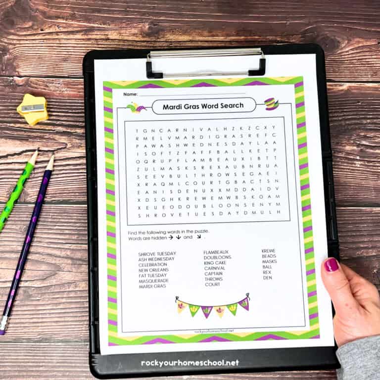Woman holding black clipboard with free printable Mardi Gras word search and green pencil, purple pencil, and yellow star-shaped pencil sharpener on wood background.
