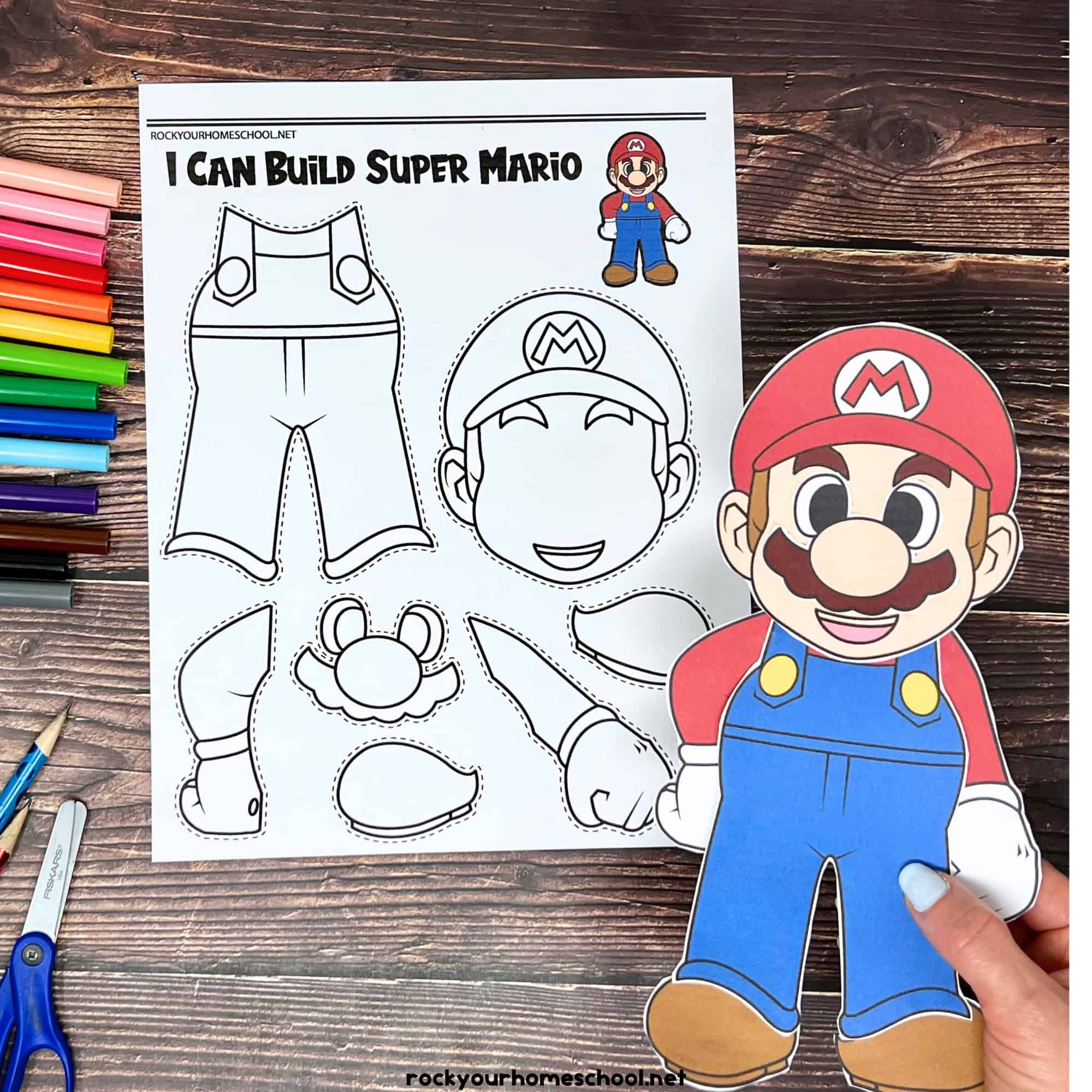 woman holding example of Super Mario craft with black-and-white version in background and markers, glue stick, pencils, and scissors.