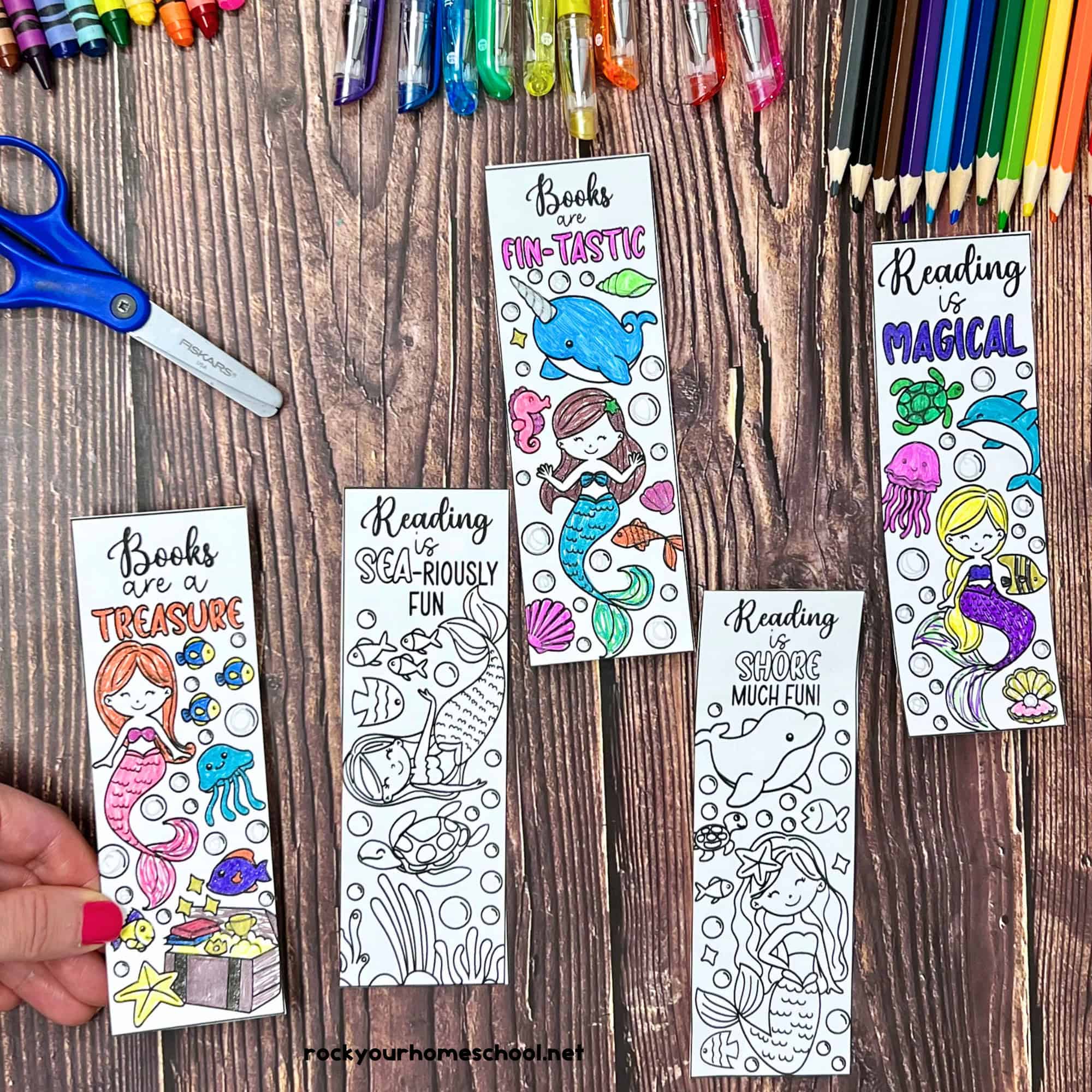 Woman holding examples of free printable mermaid bookmarks to color with crayons, glitter gel pens, color pencils, and scissors in background.