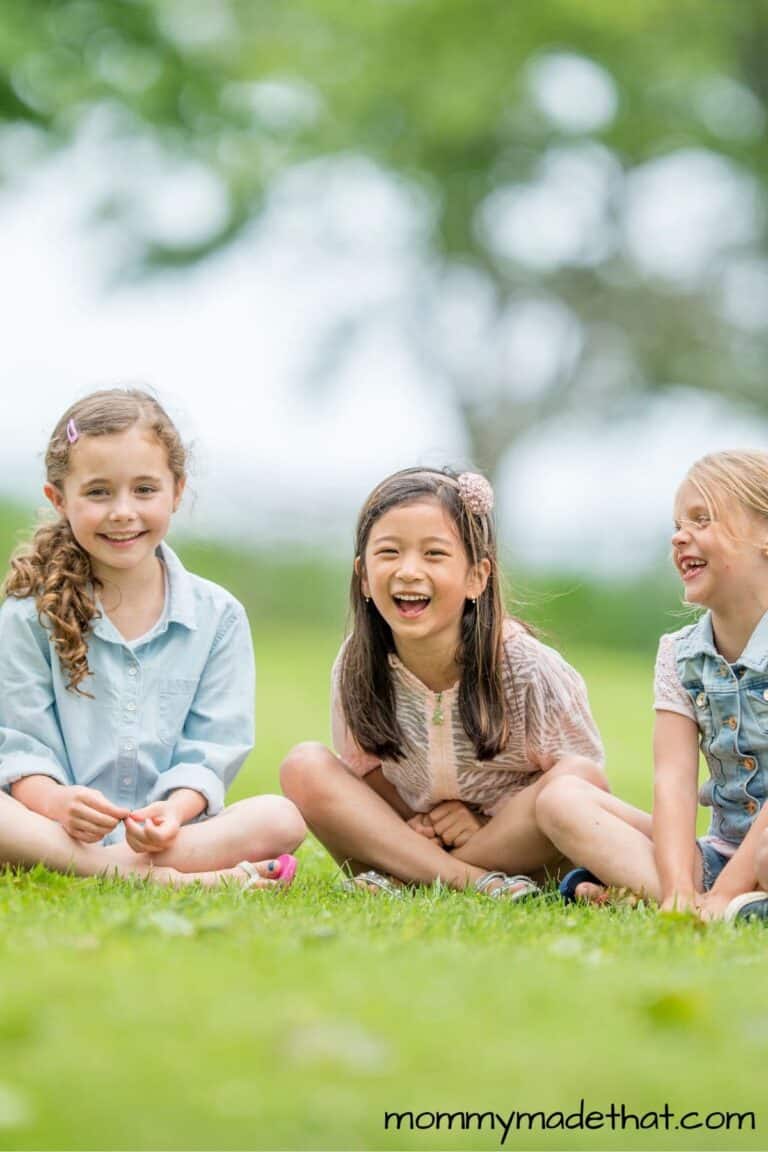 Three girls sitting down on grass as they play duck duck goose.