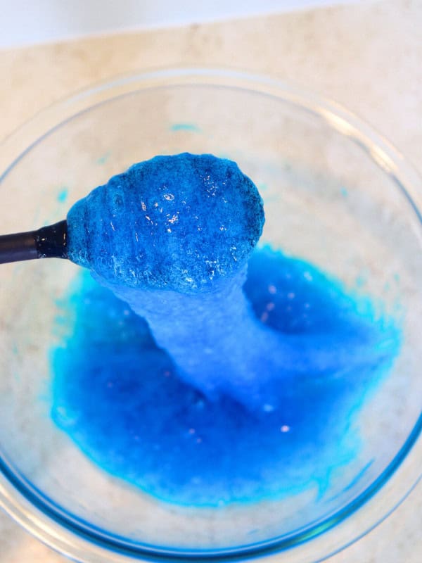 Black spoon mixing water and borax with clear glue and blue food coloring mixture.