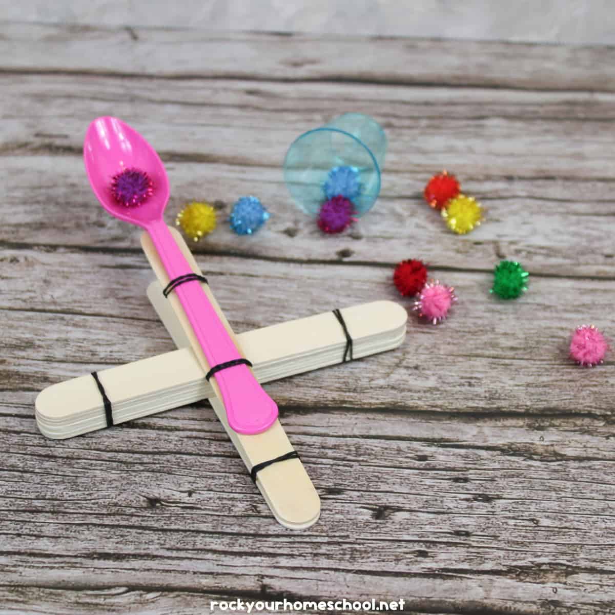 STEM catapult with wood craft sticks, rubber bands, plastic spoon, and pom poms.