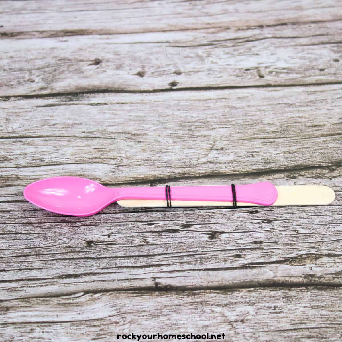 Plastic spoon on top of wood craft stick with rubber bands.