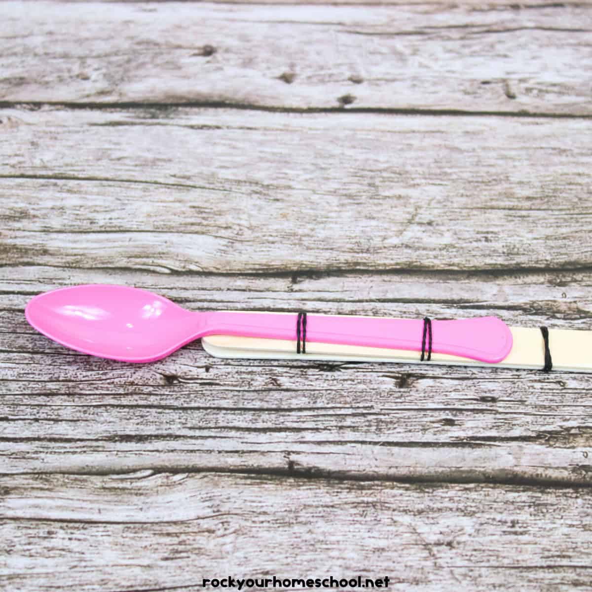 Pink plastic spoon on two wood craft sticks with rubber bans.