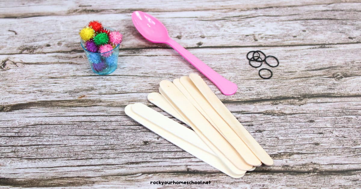 STEM catapult supplies including glitter pom poms, pink plastic spoon, rubber bands, and wood craft sticks.