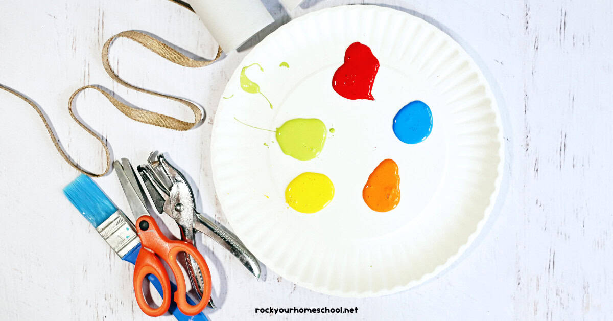 Paper plate with blue, red, orange, yellow, and green paints, ribbon, toilet paper roll, hole punch, scissors, and paint brush.