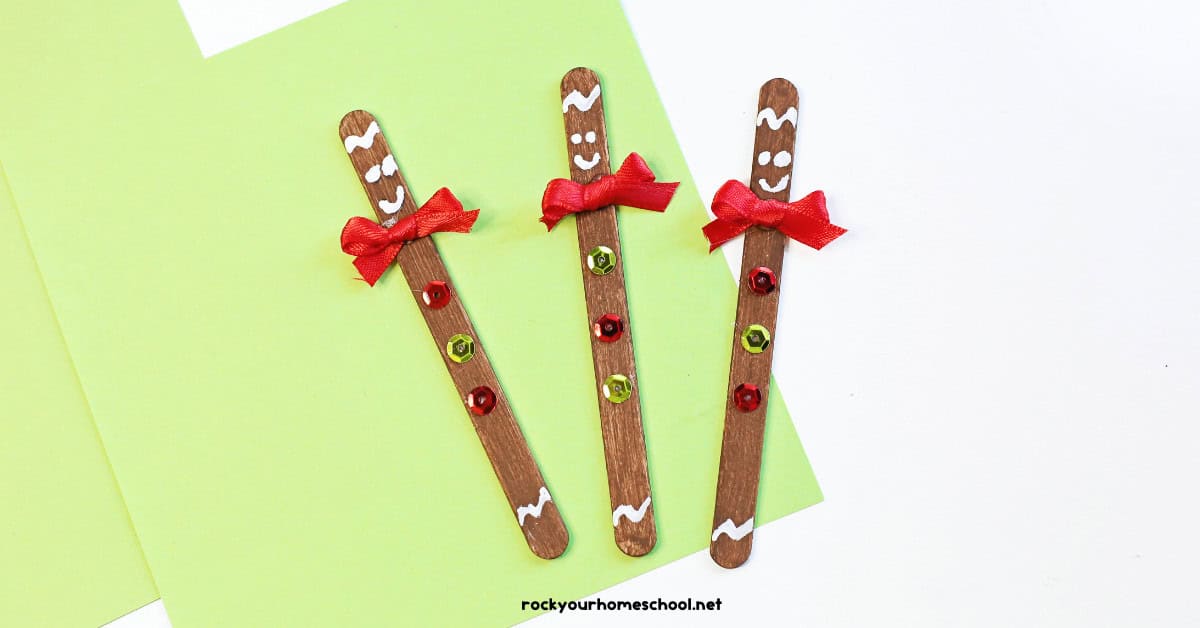 Three examples of popsicle stick gingerbread crafts with red ribbons and sequins on light green paper.