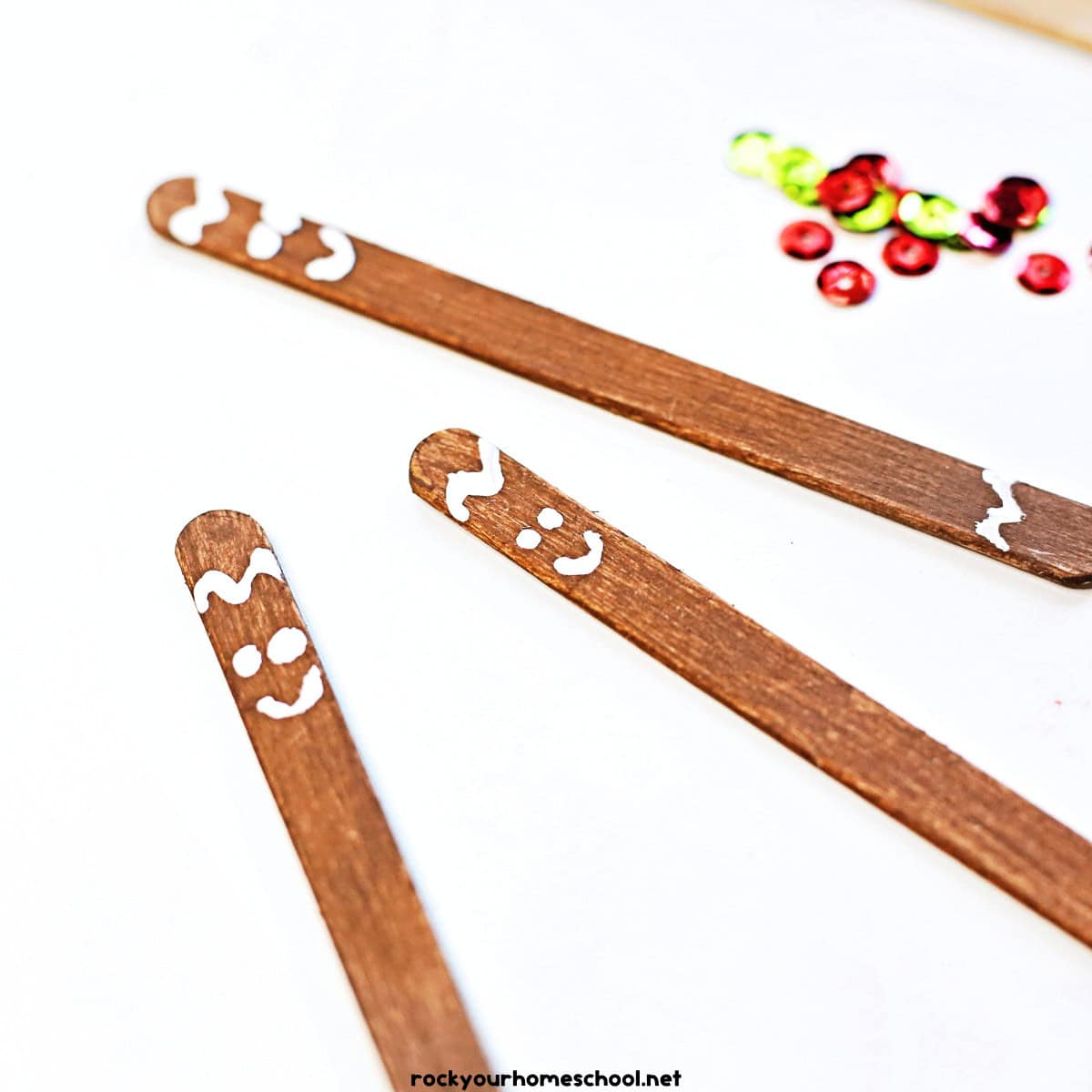 3 examples of brown popsicle sticks with white paint smiley faces and red and green sequins.