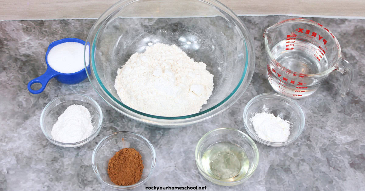 Glass bowl with flour, blue measuring cup with salt, small bowls of cream of tarter, cornstarch, cocoa powder, and vegetable oil and glass measuring cup with water.