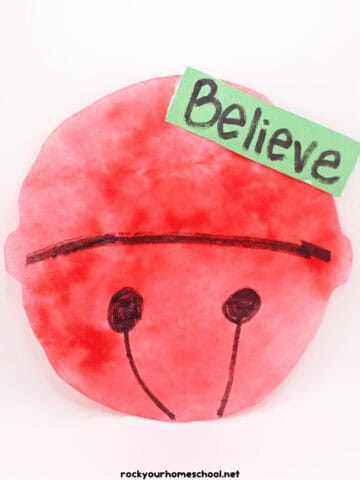 Example of red jingle bell craft made with coffee filter and markers with green paper tag that says Believe.