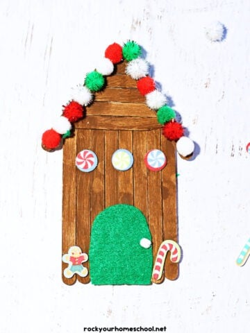 Example of popsicle stick gingerbread house with red, white, and green pom poms, foam stickers, and green felt door.