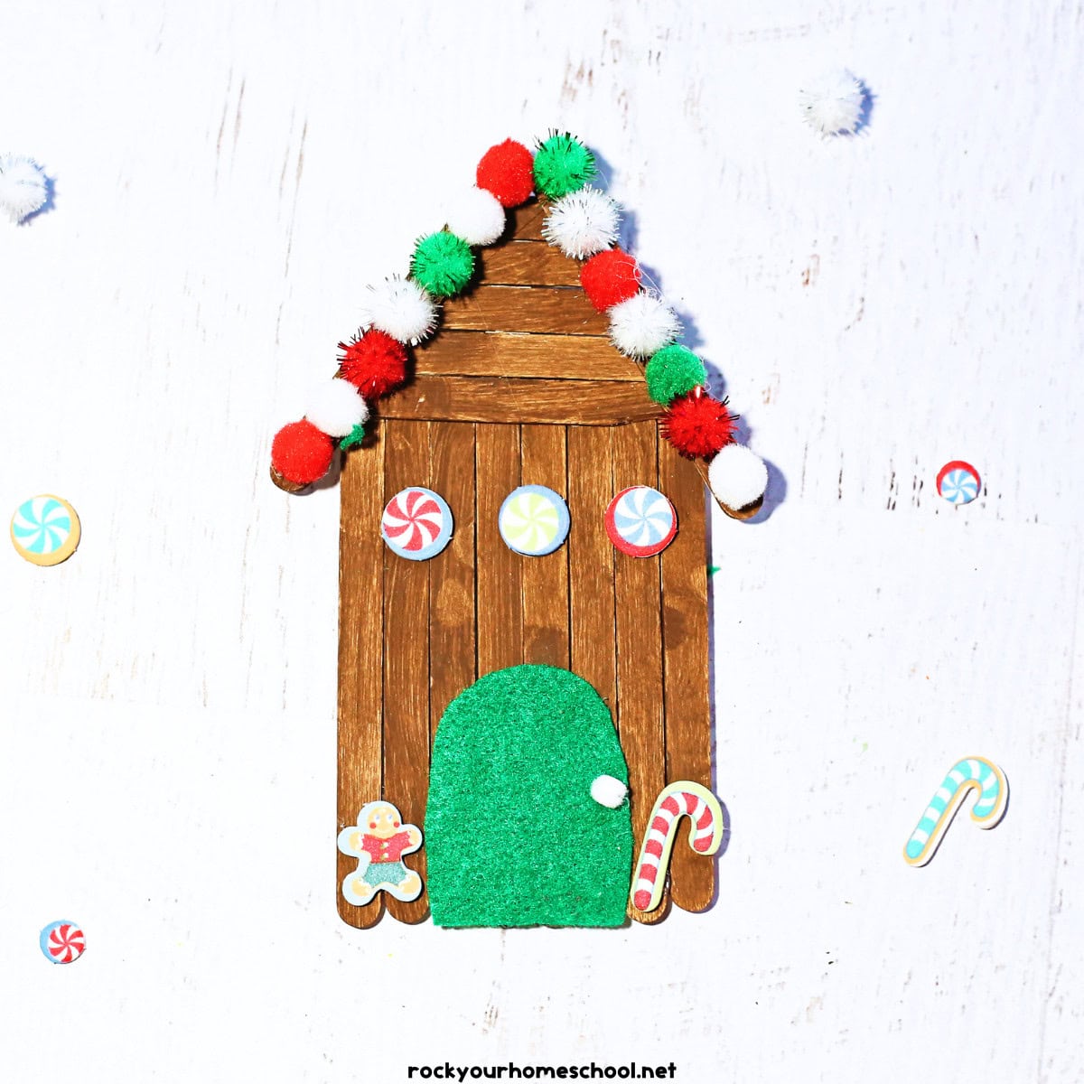 Example of popsicle stick gingerbread house with red, white, and green pom poms, foam stickers, and green felt door.