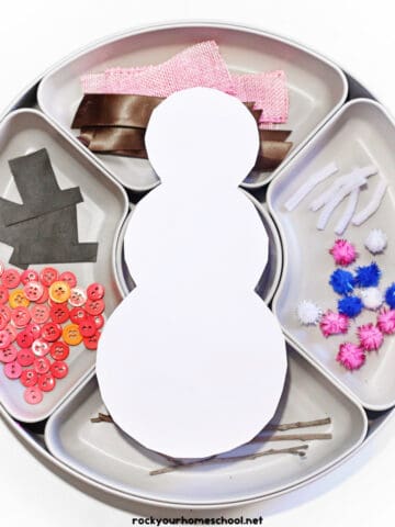 White cardstock snowman on circle tray with dividers of ribbon, pipe cleaners, pom poms, sticks, buttons, and black paper hats.