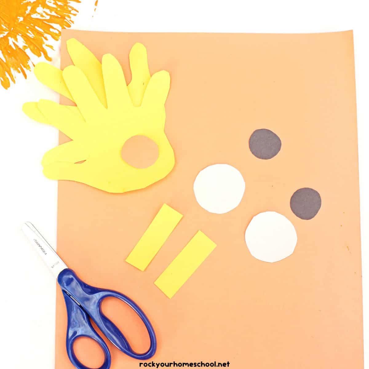 Scissors and orange paper with 2 yellow paper hands, 2 black circles, 1 white circles, and 2 yellow rectangles.