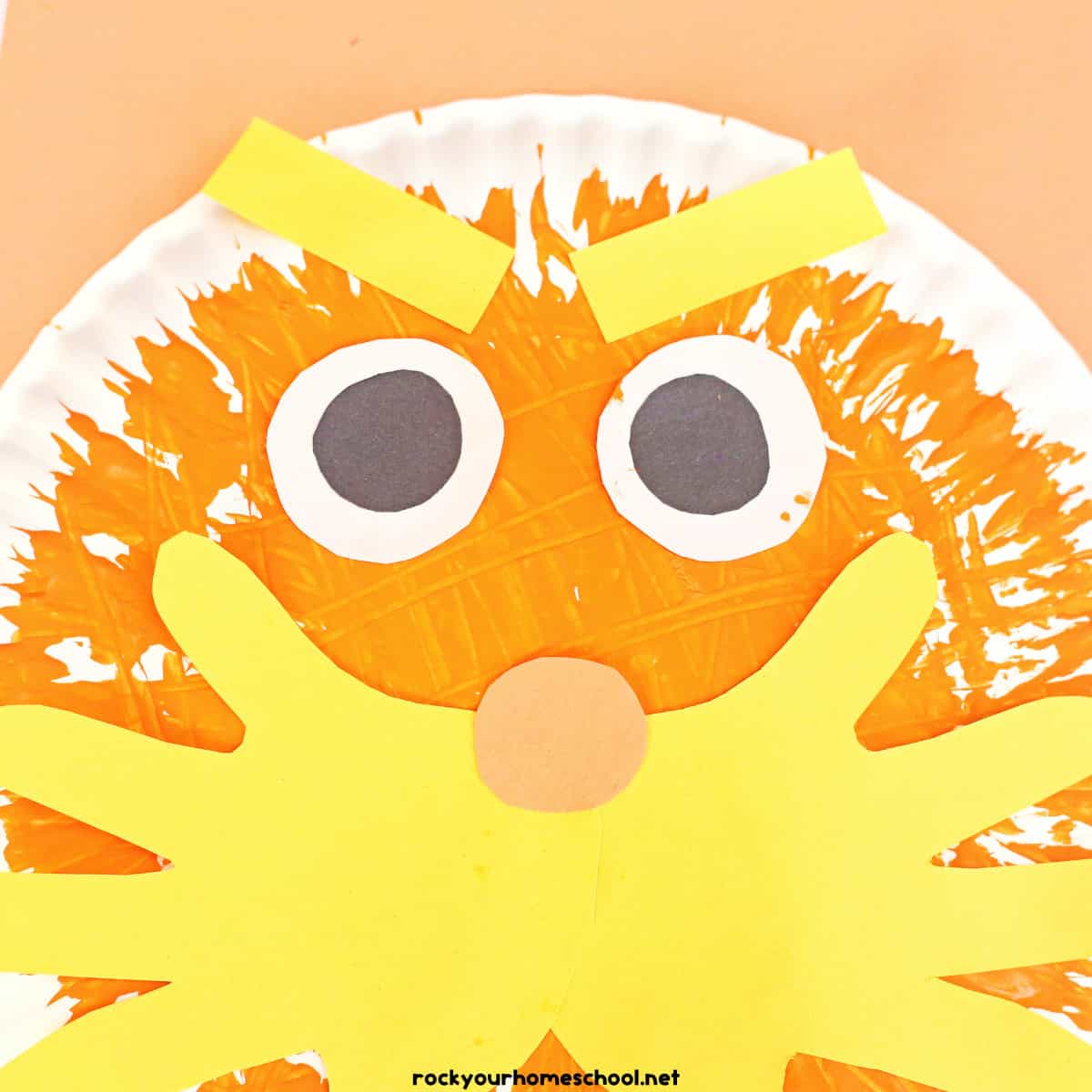 Paper plate painted orange with yellow paper handprint mustache, orange circle nose, two black and white eyes, and yellow rectangle eye brows.