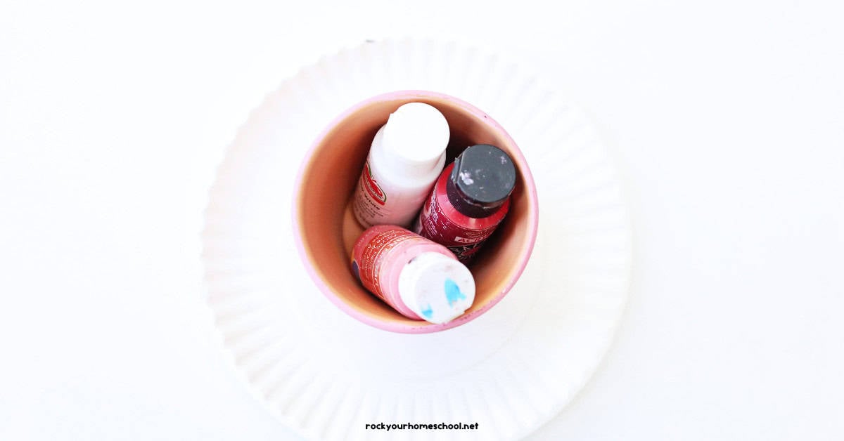 Claypot on paper plate with white, red, and pink paint bottles.