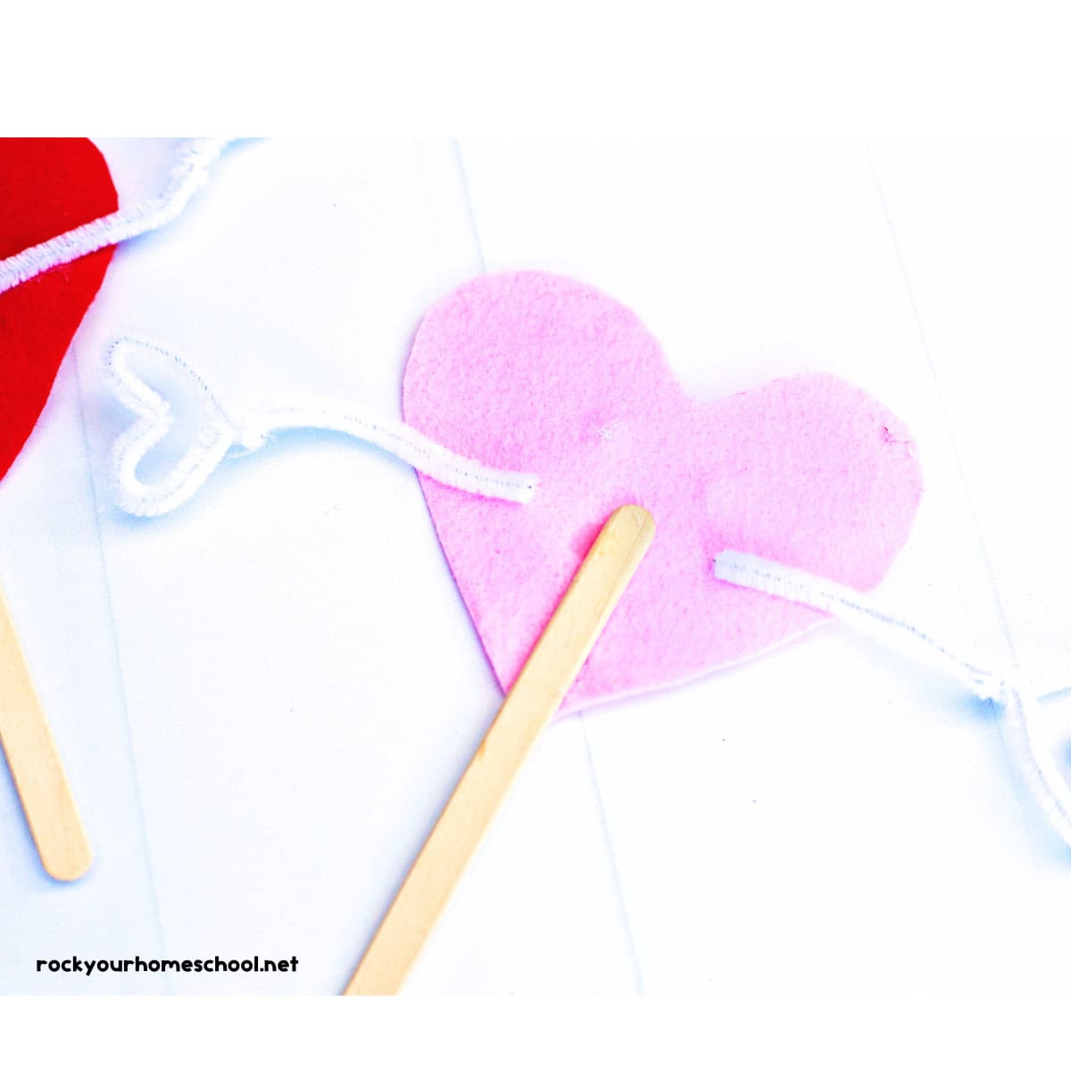 Popsicle stick glued to pink felt heart with white pipe cleaners.