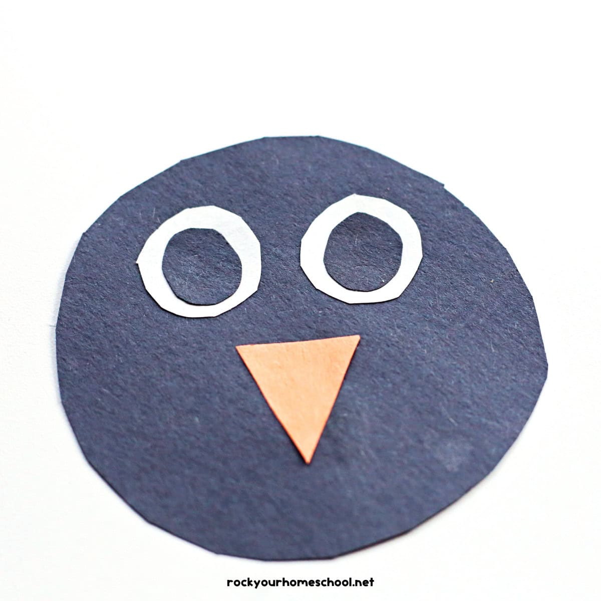 Black paper circle with white circle eyes and orange triangle beak.