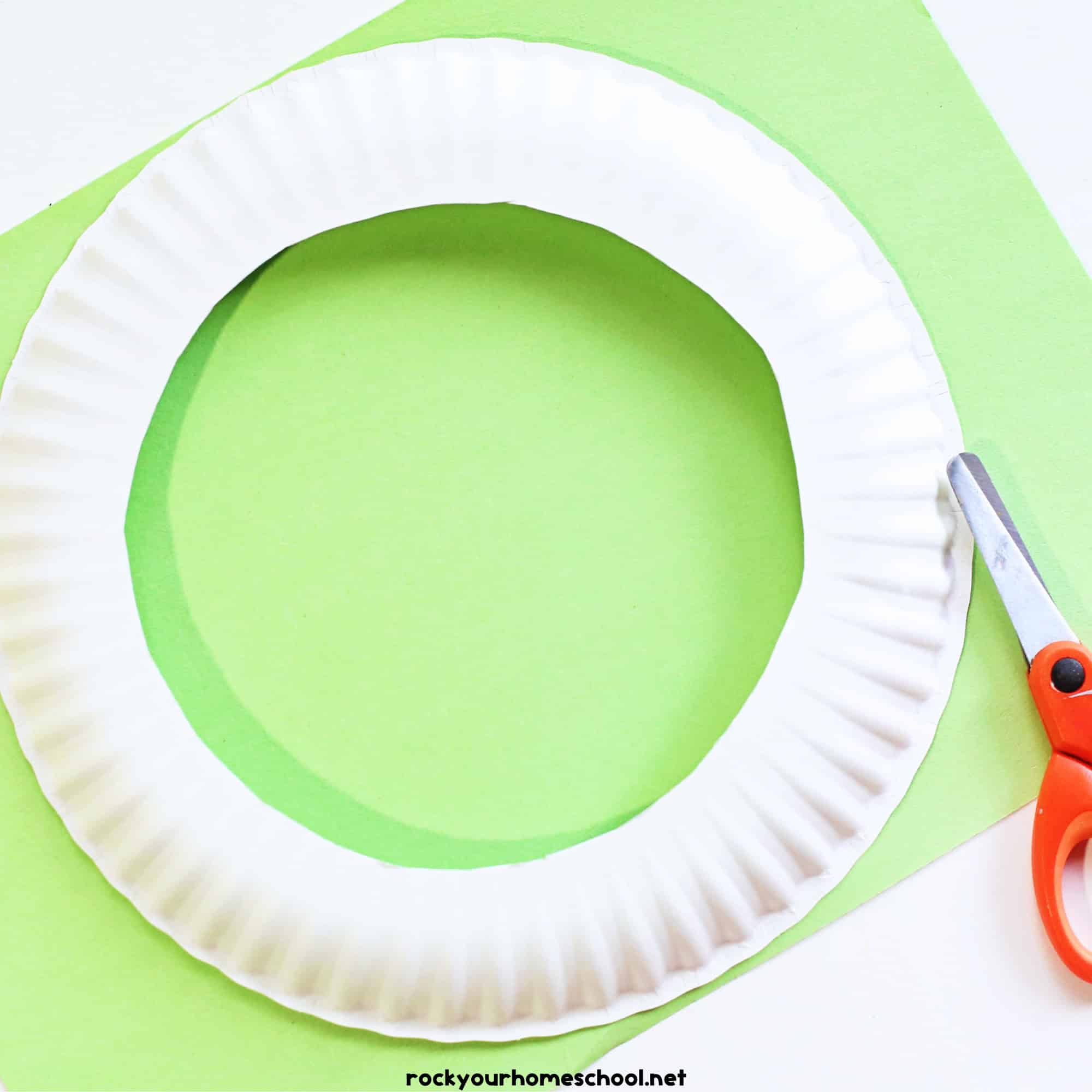 Paper plate with circle cut out of center on light green paper and scissors.