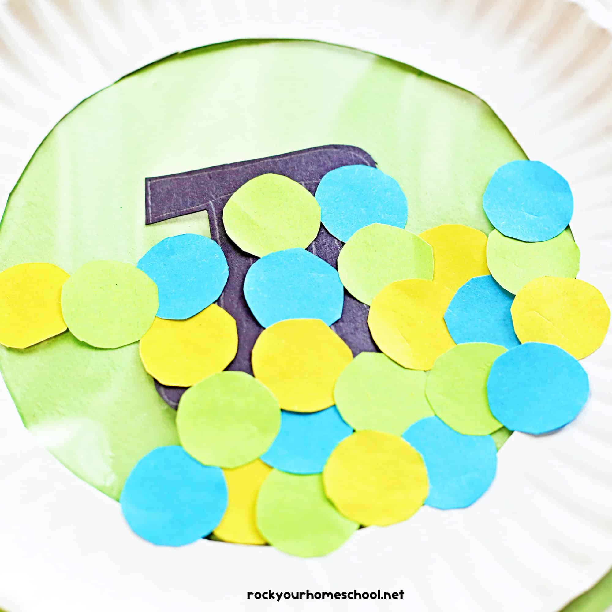 Light blue, green, and yellow paper circles on top of black paper Pi symbol and contact paper with paper plate frame.