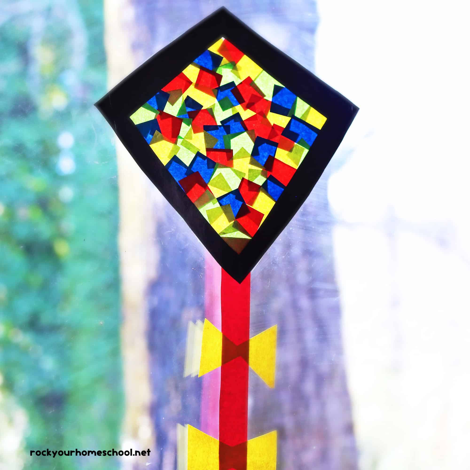 Kite suncatcher craft for kids featuring black diamond frame with red, yellow, and blue tissue paper squares with red paper string and yellow bows.