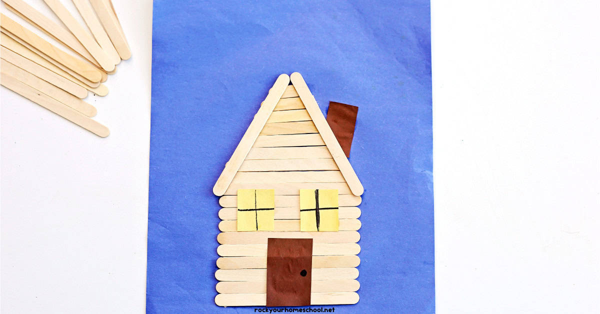 Log cabin craft on blue construction paper with popsicle sticks.