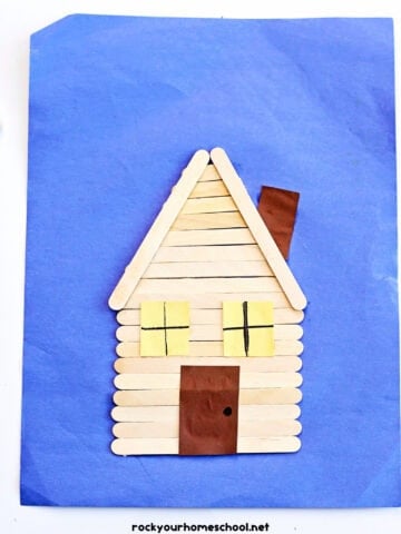 Example of popsicle stick log cabin craft on blue construction paper.