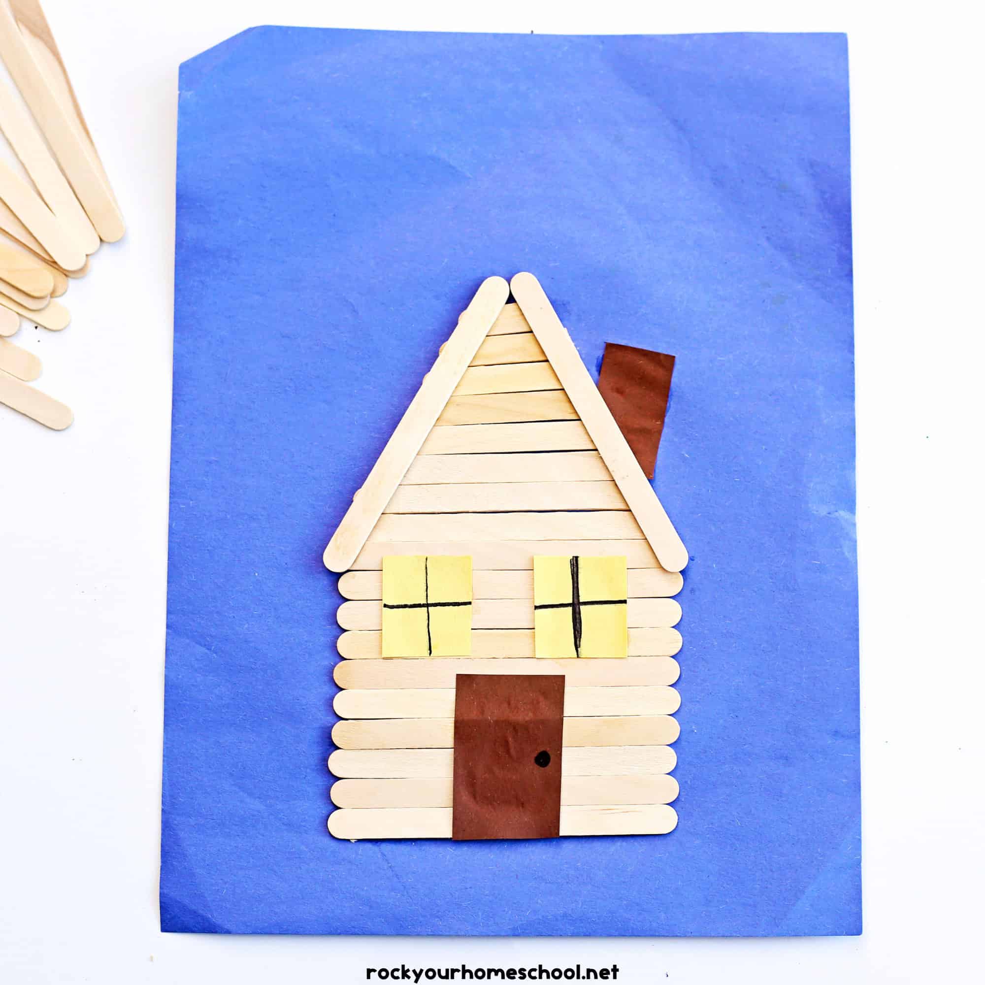 Example of popsicle stick log cabin craft on blue construction paper.