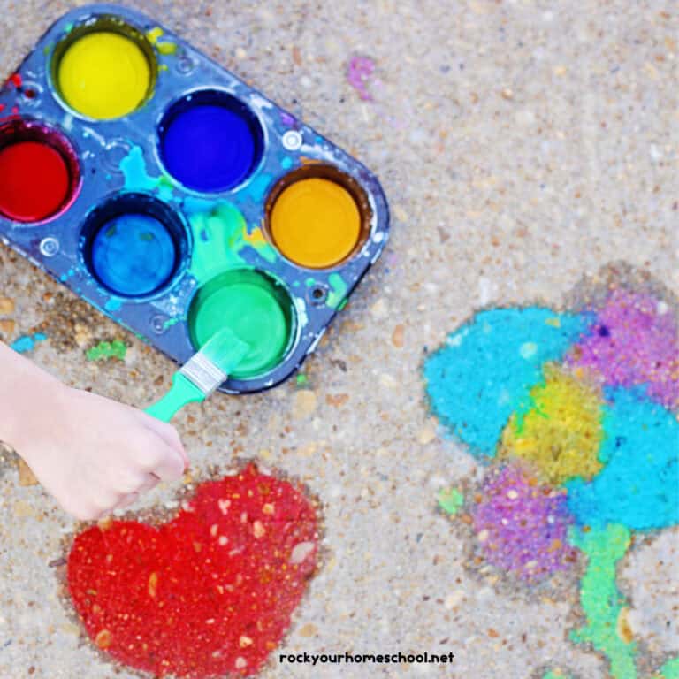 Child using green paint brush with muffin tin tray of DIY sidewalk chalk paints.