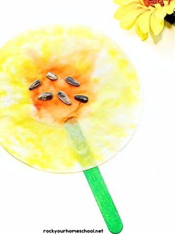 Example of coffee filter sunflower craft on green popsicle stick with sunflower seeds in center.