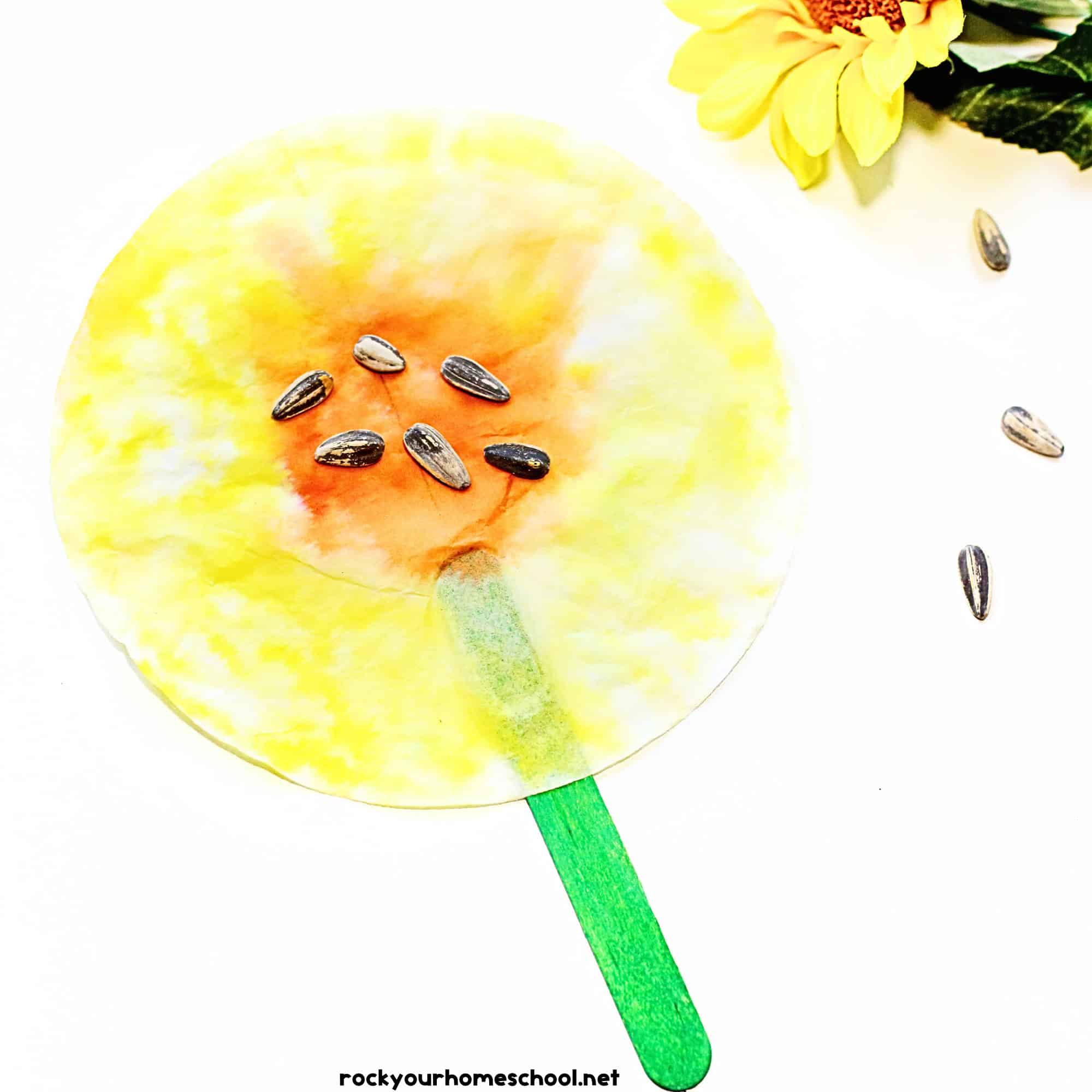 Example of coffee filter sunflower craft on green popsicle stick with sunflower seeds in center.